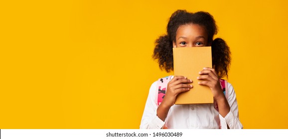 Shy African American Schoolgirl Covering Face With Book On Yellow Background In Studio. Panorama With Empty Space