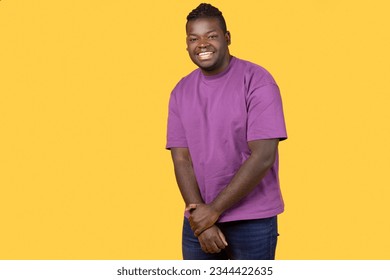 Shy African American young man looking at camera with toothy smile, holding hands together, posing wearing casual purple t-shirt, standing over yellow background, studio shot with free space - Shutterstock ID 2344422635