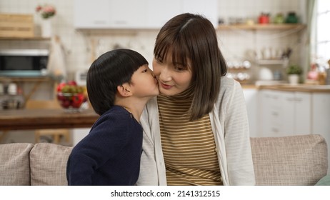 Shy Asian Little Boy Running Away After Kissing His Smiling Mother On Face In The Living Room At Home. Family And Love Concept