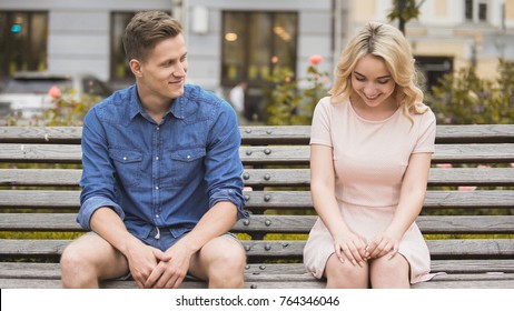 Shy blonde girl smiling, attractive guy flirting with beautiful woman on bench