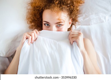 Shy Cute Lovely Young Woman With Curly Red Hair Lying In Bed And Hiding Her Face Behind Sheet