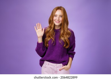 Shy And Cute Redhead Female Newbie Greeting New Team Smiling Friendly And Waving Raised Palm In Hello Or Hi Gesture, Saying Nice To Meet You As Getting Know New Member Over Violet Wall