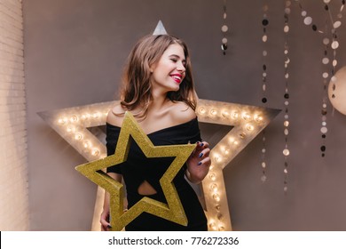 Shy girl with red lips posing in decorated studio before party. Indoor portrait of adorable lady in black holding golden star in hands.