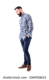 Shy handsome bearded young man in plaid shirt with hands in pockets smiling and looking down. Full body isolated on white background.