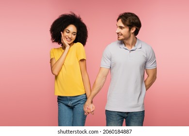 Shy Hispanic Woman Holding Hands With Smiling Man Hiding Something Behind Back Isolated On Pink