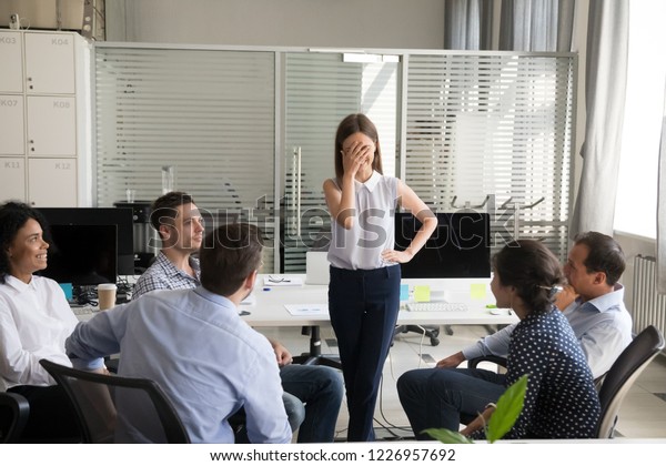 Shy nervous bashful female employee feels\
embarrassed blushing afraid of public speaking at corporate group\
team meeting, timid stressed woman hiding face during awkward\
moment reporting in office