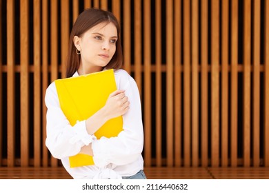 A Shy Student Girl With A Yellow Folder In Her Hands.