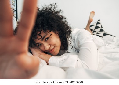 Shy Young Mixed Race Woman Looking At Camera Lying Down On The Bed Covering Lens With Hand. Selective Focus On Face.