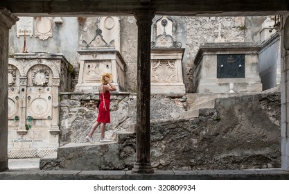 SIBENIK, CROATIA - JULY 30, 2006: A Young Woman In Red Dress Visits The The Cemetery Of The Town. Its Impressive Family Graves, Where Multiple Generations Are Buried, Attract A Lot Of Foreign Tourists