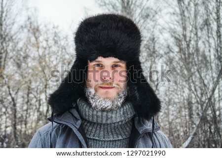 Siberian Russian man with a beard in hoarfrost in freezing cold in the winter freezes in a village in a snowdrift and wears a hat with a earflap.