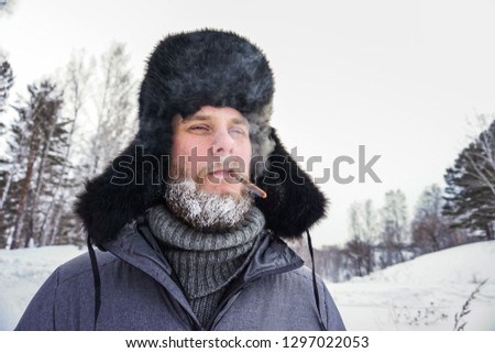 Siberian Russian man with a beard in hoarfrost in freezing cold in the winter freezes in a village in a snowdrift and wears a hat with a earflap smoking a cigarette
