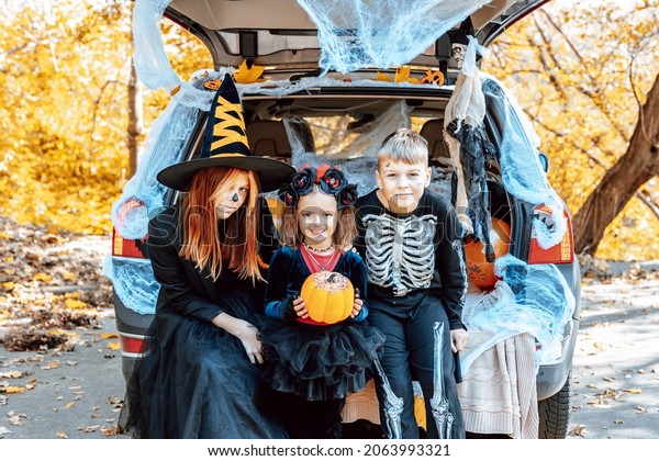siblings boy in skeleton costume, teenage girl in\
witch costume and hat and cute little girl in spooky costume sits\
in trunk car decorated for Halloween with web, orange balloons and\
pumpkins, outdoor