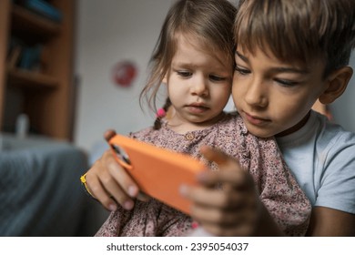 Siblings brother boy hold smartphone with girl sister children use mobile phone smartphone at home in room watch video make a call or play online games leisure family concept real people copy space - Shutterstock ID 2395054037