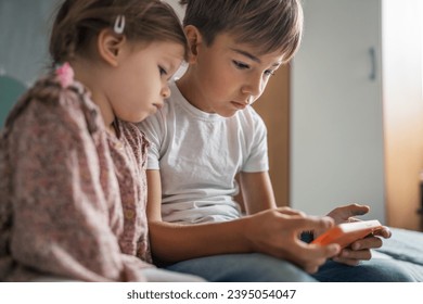 Siblings brother boy hold smartphone with girl sister children use mobile phone smartphone at home in room watch video make a call or play online games leisure family concept real people copy space - Shutterstock ID 2395054047