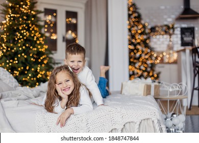Siblings brother and sister are lying in bed. Christmas morning cozy bedroom. Light Scandinavian interior. Children are cheerful happy in light sweaters. Childhood game fun. New Year European
