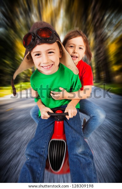Siblings in retro racing hat and\
goggles driving on toy car at speed with blurred\
background.