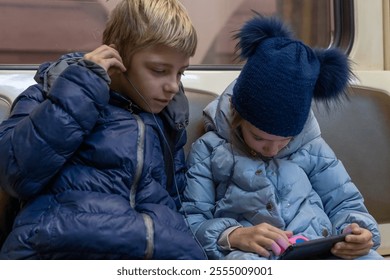 Siblings share a cozy moment on a subway, engaged with a tablet, as winter attire adds warmth to their casual outing - Powered by Shutterstock