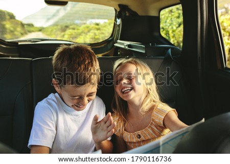 Siblings sitting on backseat of car looking at map and smiling. Kids traveling in a car on roadtrip playing with a map.