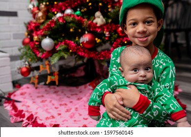 Siblings Sitting Under A Christmas Tree Wearing Matching Pajama Onesies.