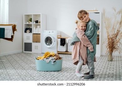 Siblings Are Standing In Middle Of The Laundry Room, Bathroom Next To A Bowl Of Clothes And Washing Machine, Children Have On Too Big Sweatshirts Hugging Each Other Tightly With Smile