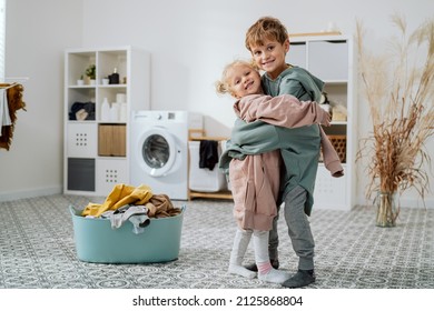 Siblings Are Standing In Middle Of The Laundry Room, Bathroom Next To A Bowl Of Clothes And Washing Machine, Children Have On Too Big Sweatshirts Hugging Each Other Tightly With Smile