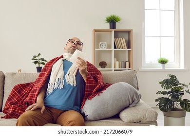 Sick adult man who has bad cold or seasonal flu virus is fighting illness at home. Unhappy middle aged man is sitting on sofa wrapped in warm scarf and plaid, holding paper tissue and sneezing