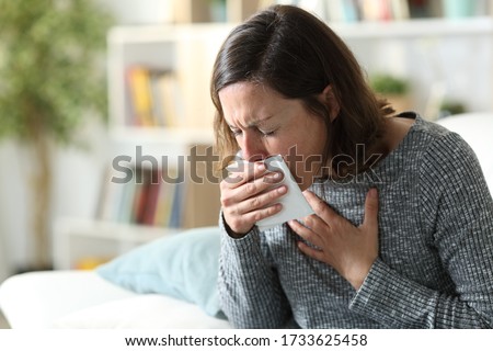 Sick adult woman coughing covering mouth with tissue sitting on a couch at home