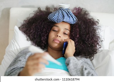 Sick African American Girl With Flu In Bedroom At Home. Ill Young Black Woman With Cold Calling Doctor By Phone, Holding A Thermometer In Her Mouth.