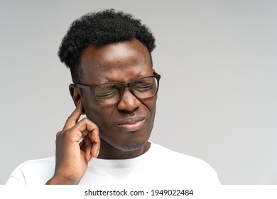 Sick African American millennial man suffering from tinnitus, throbbing earache, tired of noise. Irritated black frowning male in glasses touching painful ear, isolated on grey studio background.