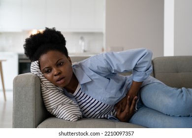 Sick African American Woman Experiencing Health Problems Suffering From Cramps And Stomach Pain. Young Black Girl Rubs Belly With Hands During Menstruation, Feels Ache Lies On Sofa In Living Room 