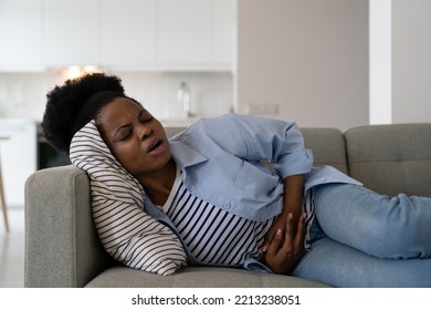 Sick African American Woman Experiencing Health Problems Suffering From Cramps And Stomach Pain. Young Black Girl Rubs Belly With Hands During Menstruation, Feels Ache Lies On Sofa In Living Room 