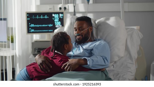 Sick african man lying in hospital bed welcoming and hugging little daughter visiting. Cute afro-american girl visit and embrace ill father in intensive care unit