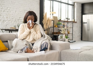Sick African-American woman sitting covered with blanket on the couch and blowing nose in paper tissue, suffering from seasonal allergy of viral disease, female with runny nose using handkerchief