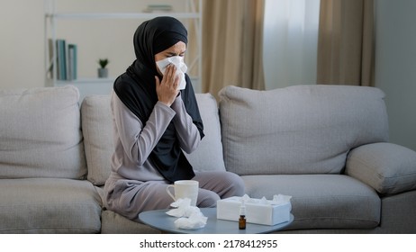 Sick arabian muslim young unhealthy woman in hijab suffer from runny nose flu disease seasonal allergy sneeze blow nose in paper napkin symptoms of coronavirus drink hot tea sit at home self-isolation