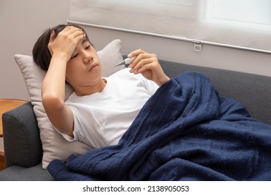 Sick Asian bot teen with thermometer lying on sofa with blue blanket.