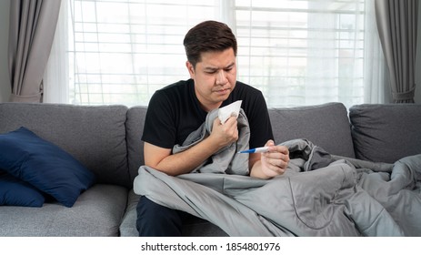 Sick Asian Man With Flu Virus Lying In Sofa With High Fever And Measuring Body Temperature By Thermometer And Touching His Forehead At Home