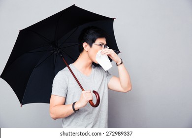 Sick Asian Man Holding Umbrella And Blowing Nose Over Gray Background