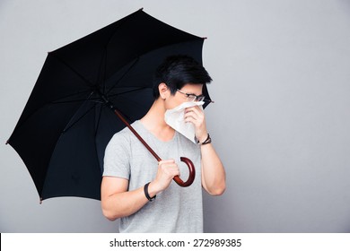 Sick Asian Man Holding Umbrella And Blowing Nose Over Gray Background