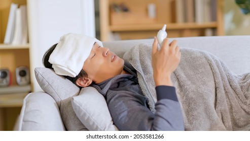 Sick Asian Man Lying On Couch Gets A Fever And Puts Towel On Forehead To Cooling Down His Body Temperature In Living Room At Home - He Measures Body Temperature With Ear Thermometer