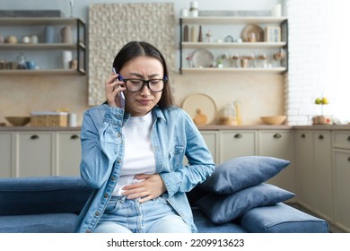 Sick Asian Woman At Home, Woman Sitting On Sofa And Having Severe Stomach Pain, Calling Hospital For Help, Young Girl Alone At Home In Kitchen