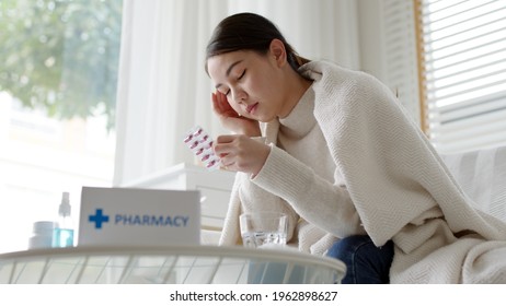 Sick Asian Woman Sit Under Blanket Acute Severe Headache On Sofa At Home. Female Hold Or Check Drug Medicine Capsule At Home, Suffer From Flu. Cold Or Fever Side Effect After Covid19 Vaccine Shot.