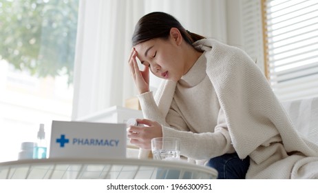 Sick Asian Woman Sit Under Blanket Acute Severe Headache On Sofa At Home. Female Hold Or Check Drug Medicine Capsule At Home, Suffer From Flu. Cold Or Fever Side Effect After Covid19 Vaccine Shot.