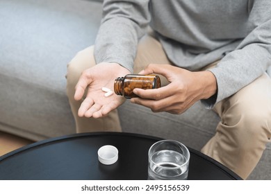 Sick asian young man, male holding tablet pill on hand, painkiller medicine from fever, stomach pain, headache for treatment, take drug or vitamin with glass of water on at home. Health care concept. - Powered by Shutterstock