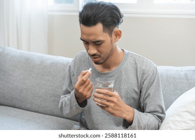 Hombre joven asiático enfermo, hombre que sostiene la píldora de la tableta en la mano, medicina analgésica de la fiebre, dolor de estómago, dolor de cabeza para el tratamiento, toma la droga o la vitamina con el vaso de agua en casa. Concepto de atención de salud.