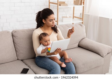 Sick Baby. Worried Black Mom Caring For Ill Toddler Son With Fever, Using Tablet Browsing Symptoms And Holding Thermometer Sitting On Sofa At Home. Online Medical Treatment Concept