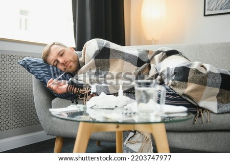 Sick bearded man who has bad cold or seasonal flu sitting on couch at home. Guy with fever wearing warm plaid shivering with worried face expression