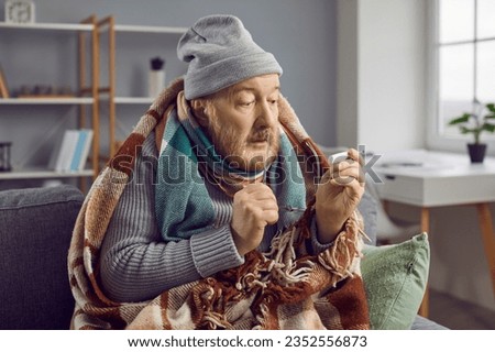 Sick bearded senior man with very high temperature sitting in blanket and hat on couch at home and looking with surprised face expression at thermometer in his hand. Sickness, cold, flu, fever concept