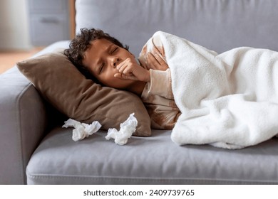 Sick black boy feeling unwell at home, coughing into fist while lying on couch under white blanket with tissues nearby, ill male kid resting during the day in quiet living room, closeup - Powered by Shutterstock