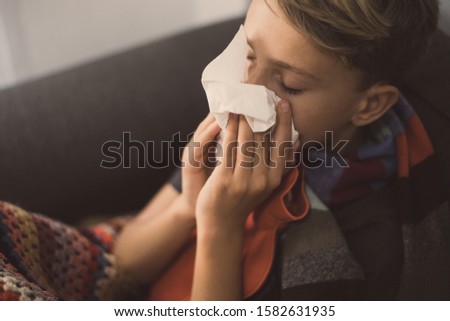 Sick boy with cold rhinitis sits with woolen blanket and hot water bottle. Sad teen with the flu blow his nose with napkin. Child with seasonal infections and fever Health disease and drugs concept