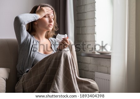 Sick brunette woman with headache having cold and flu sneezing sitting on the couch in the living room near window